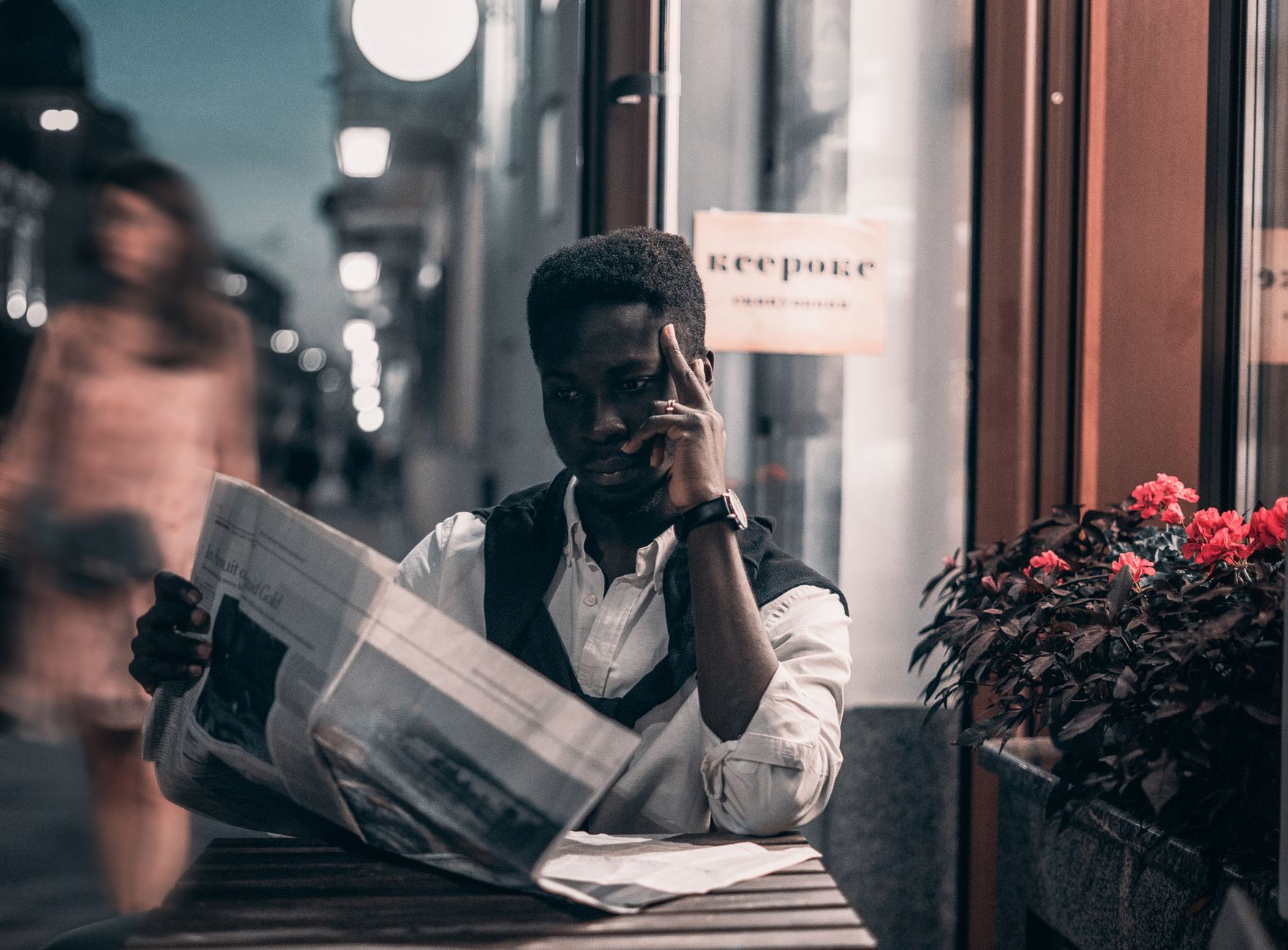 A fashionable man reads a newspaper at an outdoor cafe in the city at twilight, adding a touch of style to the urban scene. —
