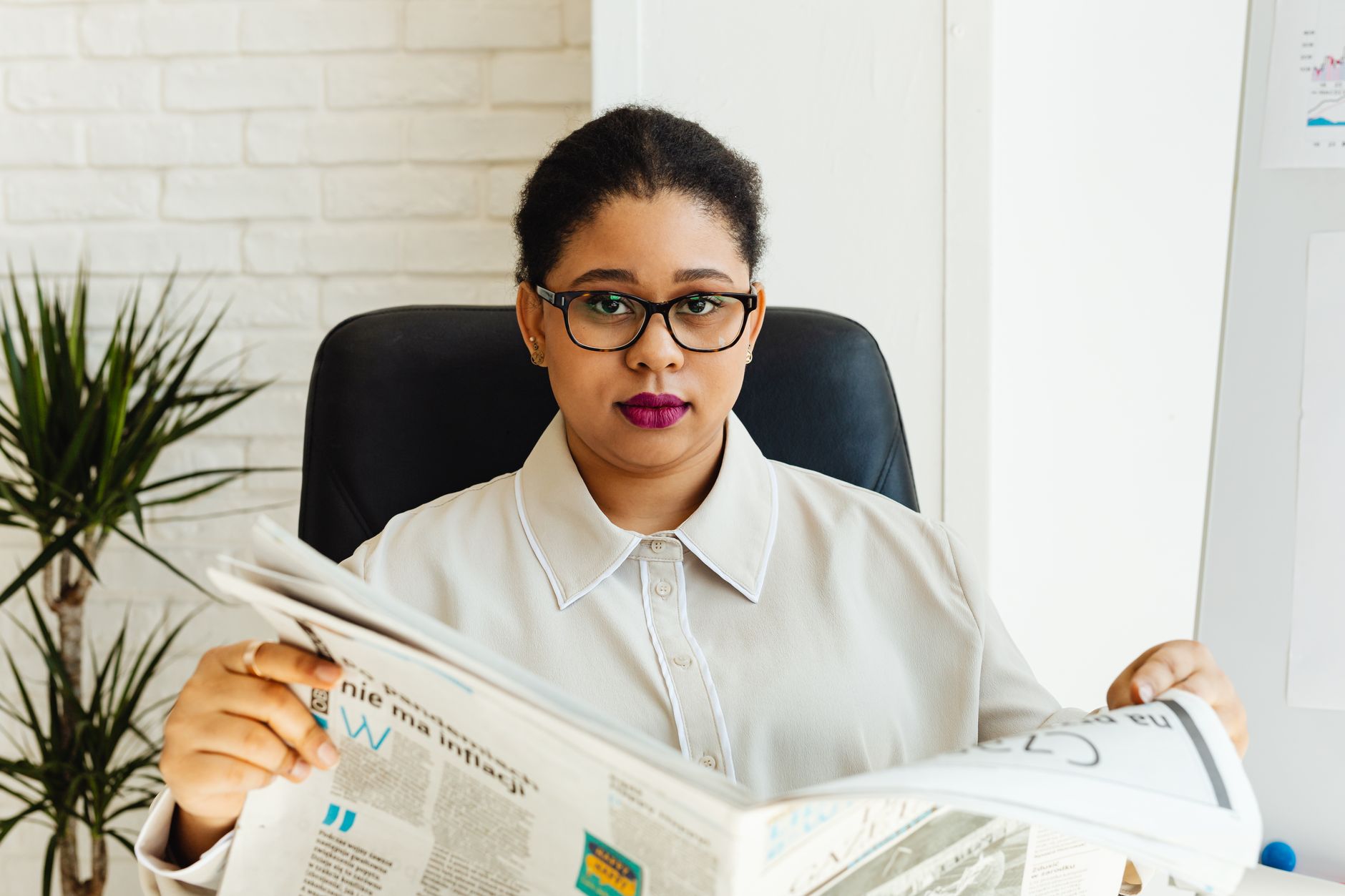 Professional woman with glasses reading a newspaper at her office desk, surrounded by modern decor. — Vavada 30 com
